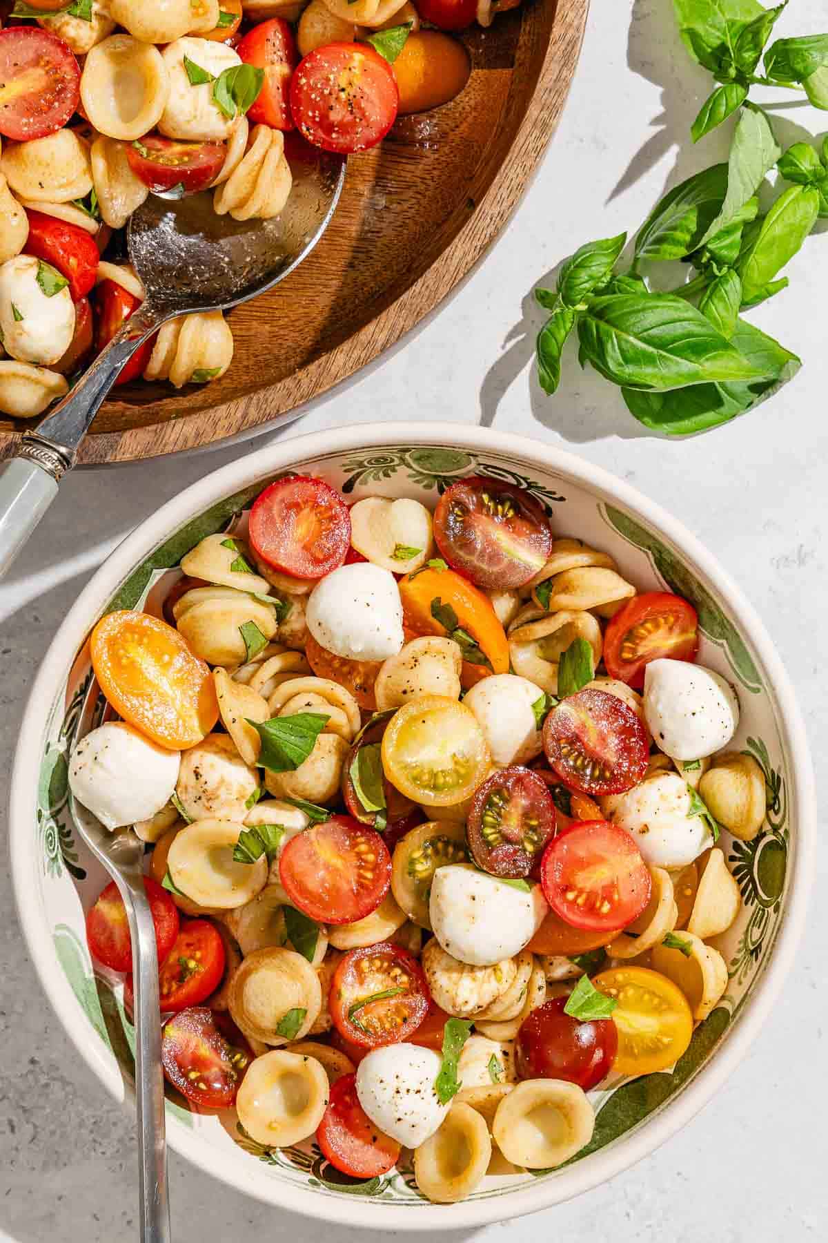 A serving of caprese salad in a bowl with a fork next to the rest of the salad in a serving bowl with a spoon and some fresh basil leaves.
