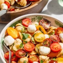 A serving of caprese salad in a bowl with a fork next to the rest of the salad in a serving bowl with a spoon.