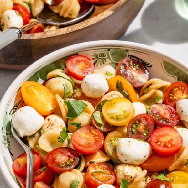 A serving of caprese salad in a bowl with a fork next to the rest of the salad in a serving bowl with a spoon.