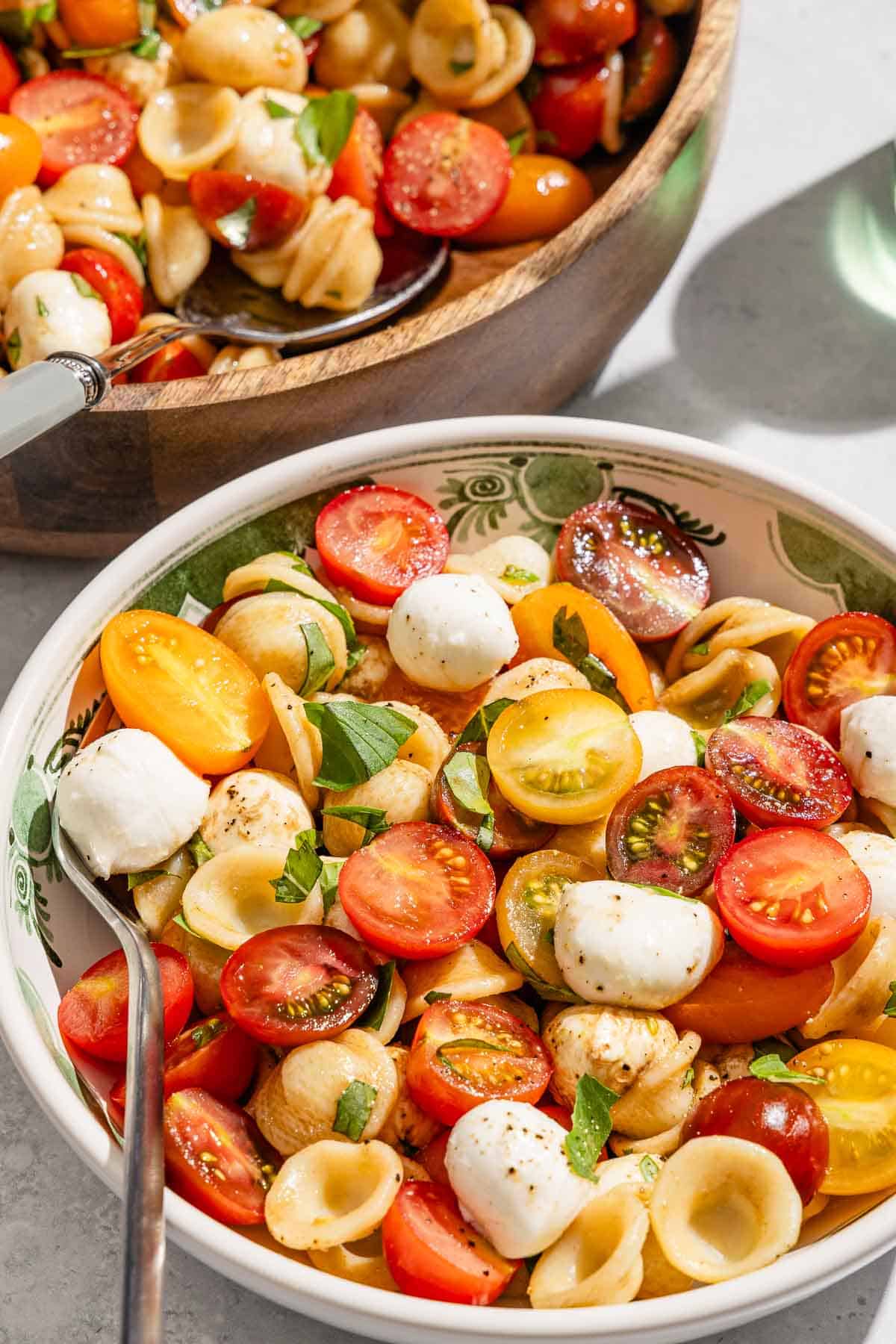 A portion of Caprese salad in a bowl with a fork next to the rest of the exhausted in a serving bowl with a spoon.