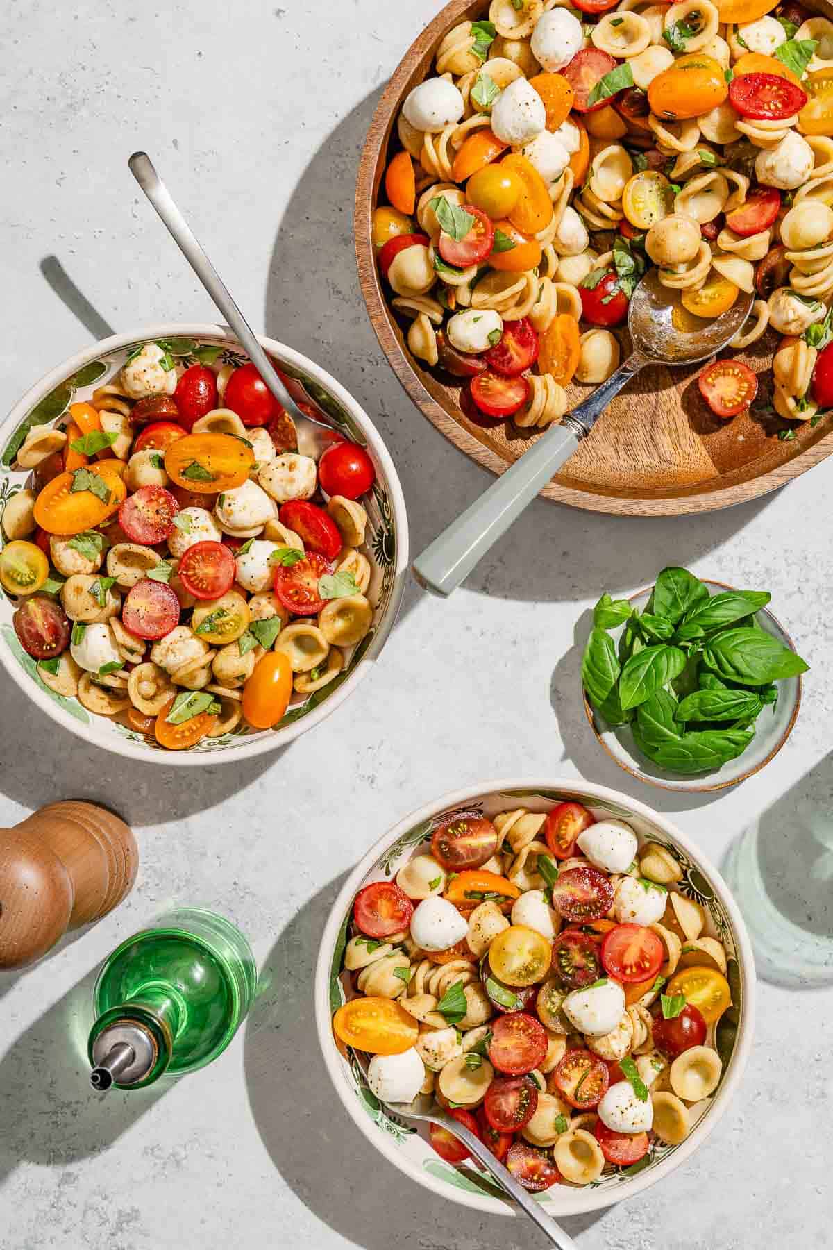 Two portions of caprese pasta salad in bowls with forks. Next to these there is a serving bowl with the rest of the salad, a small bowl of basil, a bottle of olive oil and a chopped grinder.
