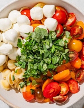 The ingredients for the caprese pasta salad I a mixing bowl just before being mixed together. Next to this is a bottle of olive oil, and bowl of balsamic vinegar, and a wooden spoon.
