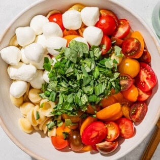 The ingredients for the caprese pasta salad I a mixing bowl just before being mixed together. Next to this is a bottle of olive oil, and bowl of balsamic vinegar, and a wooden spoon.