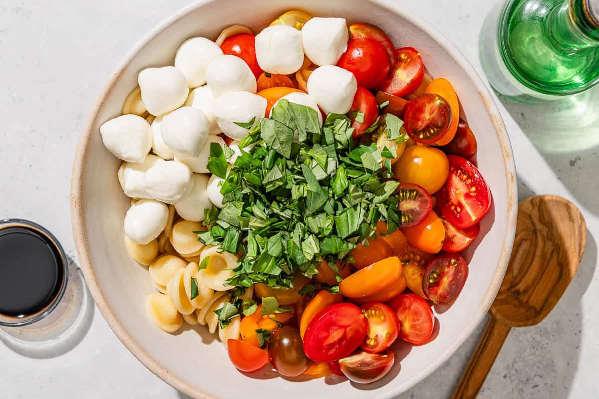 The ingredients for the caprese pasta salad I a mixing bowl just before being mixed together. Next to this is a bottle of olive oil, and bowl of balsamic vinegar, and a wooden spoon.
