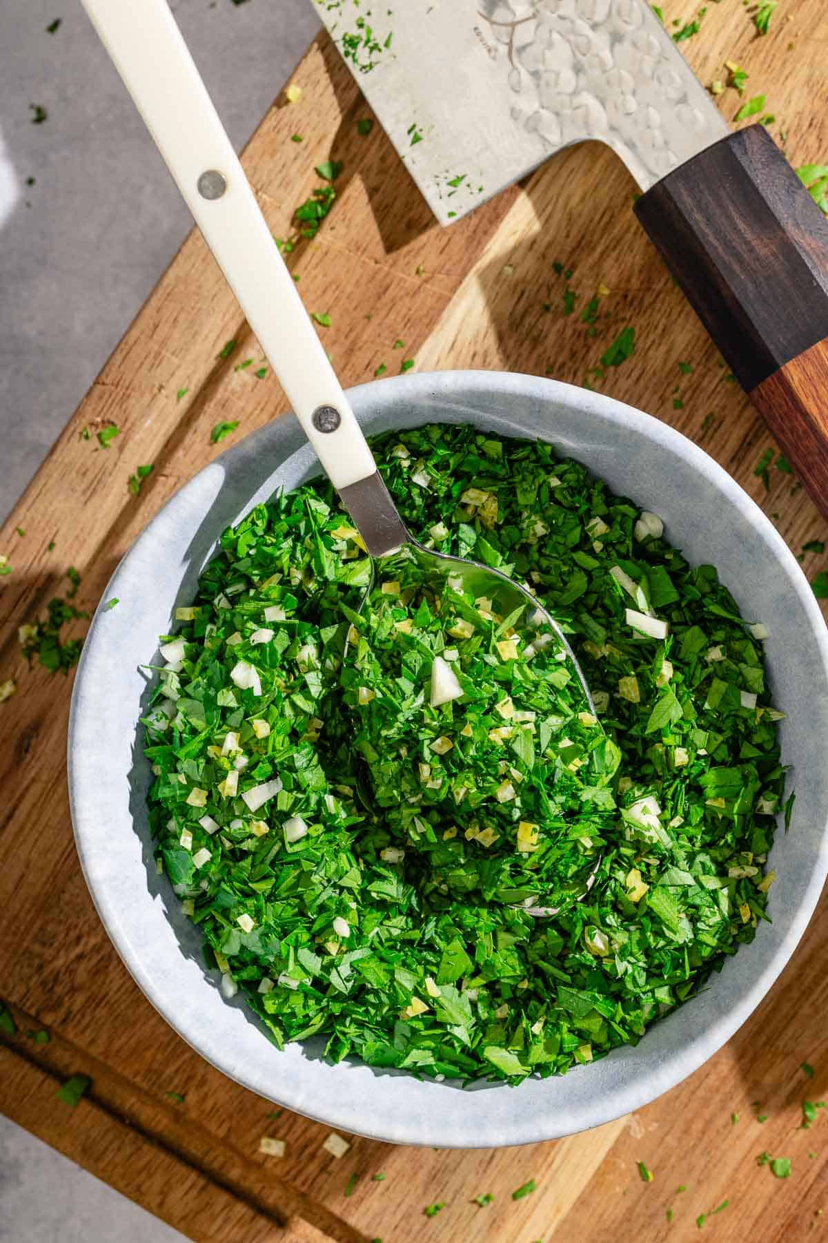 Gremolata in a bowl with a spoon next to a knife on a cutting board.