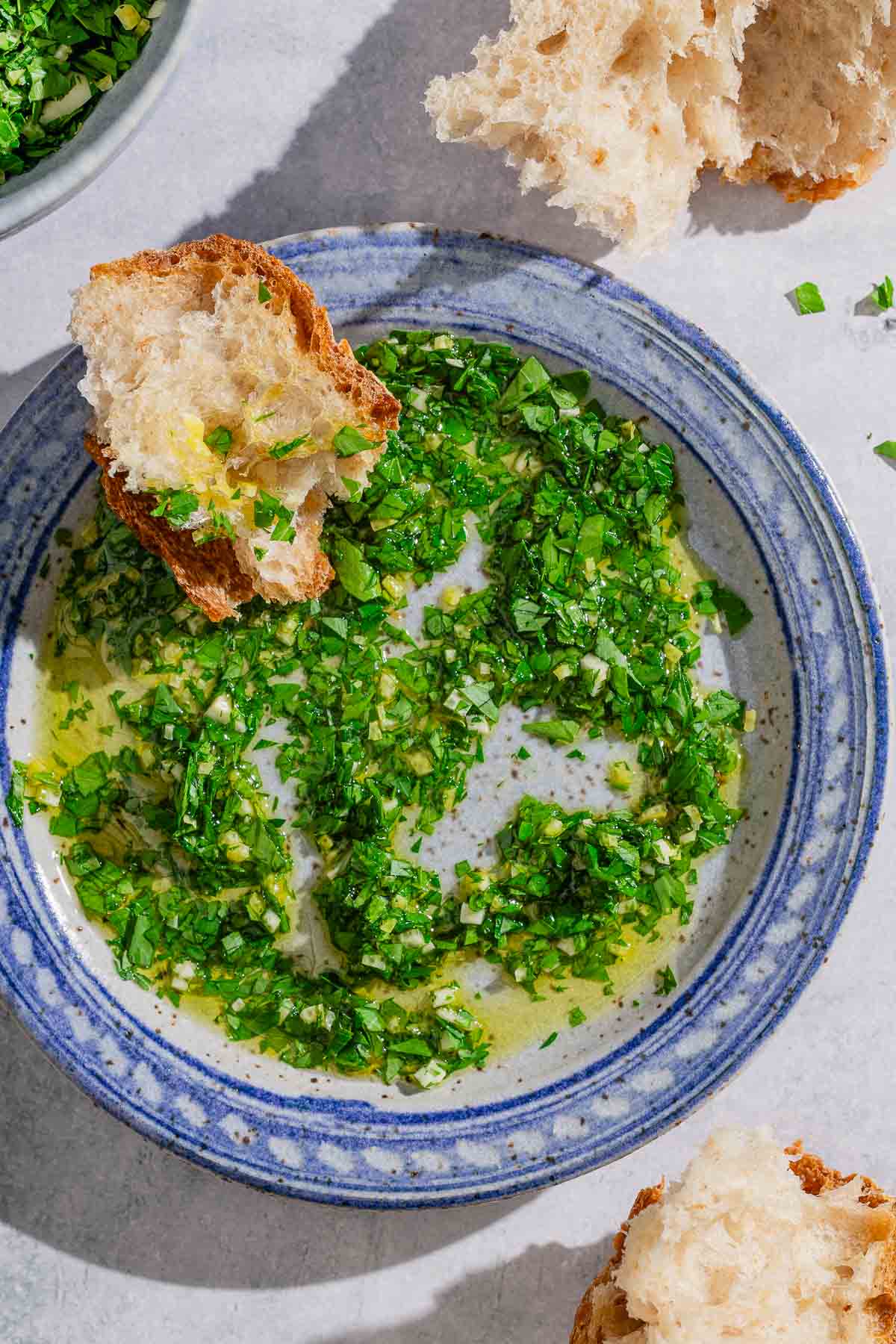 Gremolata mixed with olive oil on a plat with a slice of bread. Next to this are more pieces of crusty bread.