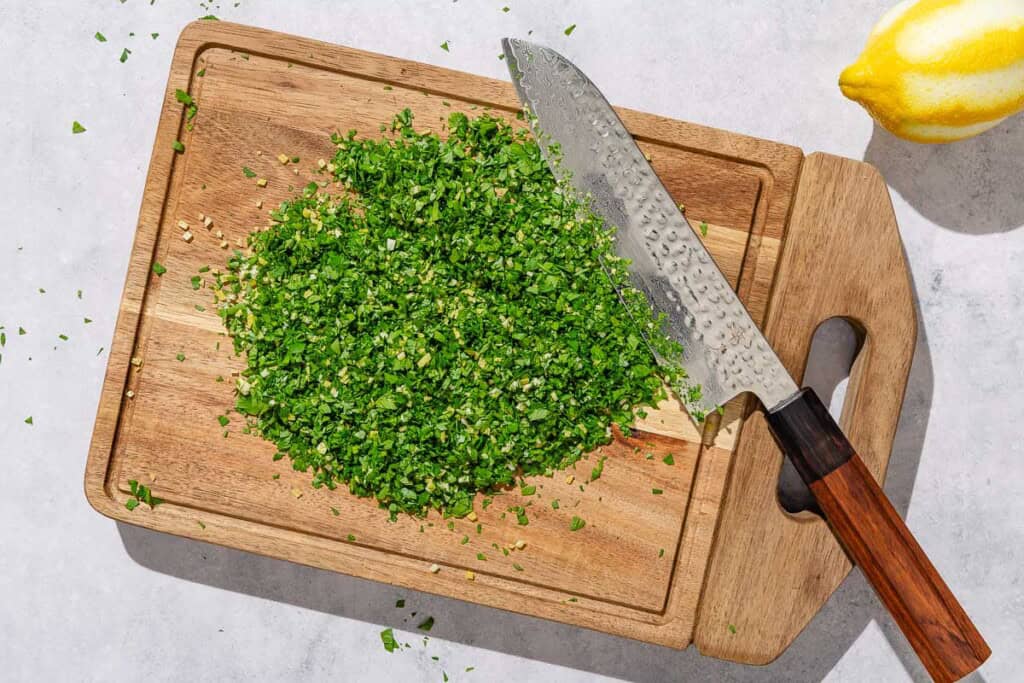 Gremolata on a cutting board with a knife. Next to this is a zested lemon.