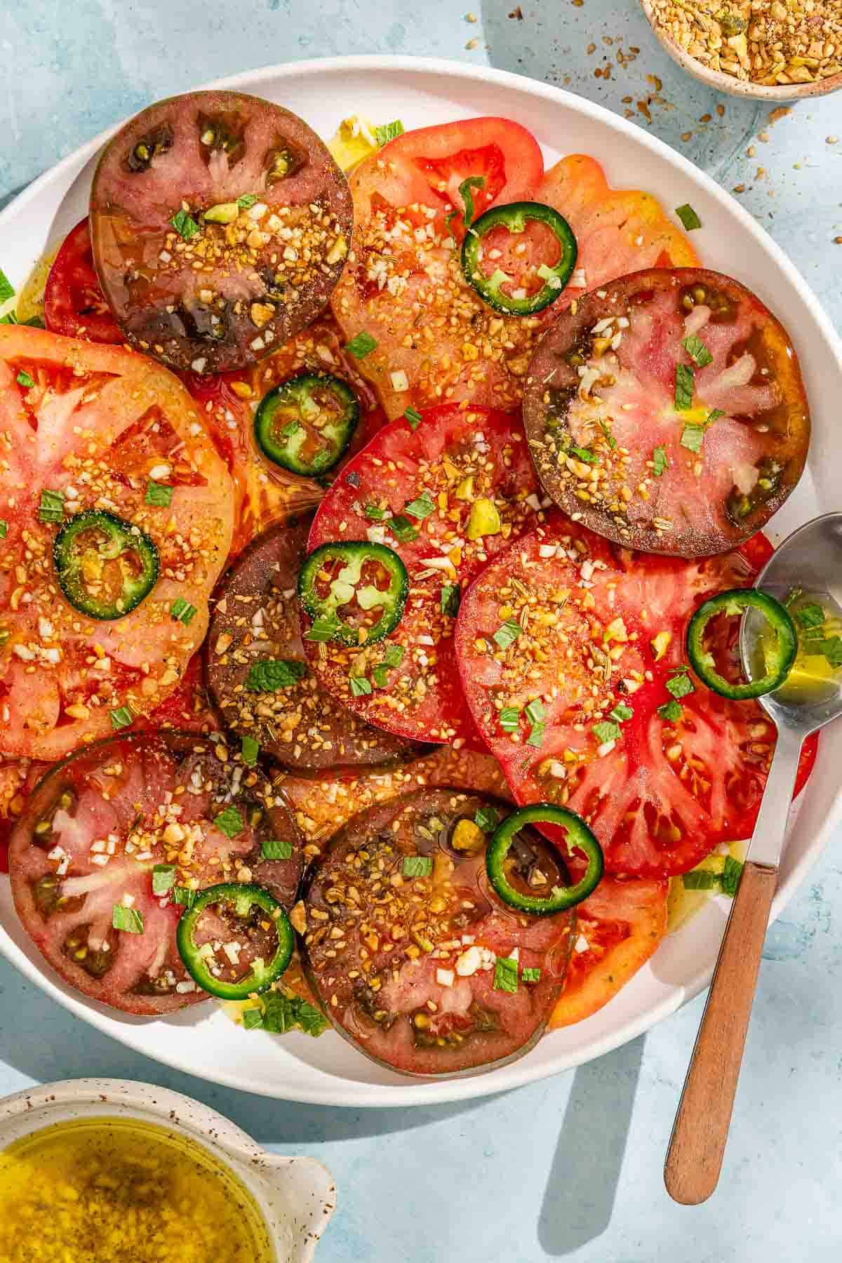 Heirloom tomato salad topped with dukkah and mint on a platter with a spoon next to a cup of the dressing and a small bowl of dukkah.