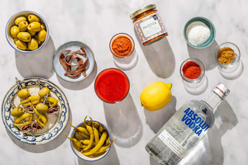 Ingredients for Mediterranean bloody Marys including tomato juice, lemon juice, olives, vodka, harissa paste, kosher salt, smoked paprika, ground cumin, lemon, pickled peppers and anchovies.