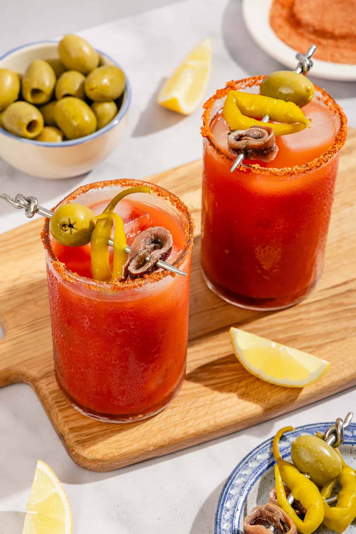 Two Mediterranean bloody Marys garnished skewers of olives, pickled peppers and anchovies on a wooden cutting board. Next to these is a bowl of green olives, scattered lemon wedges, a plate of skewered garnishes, and a plate of the spices used to rim the glasses.