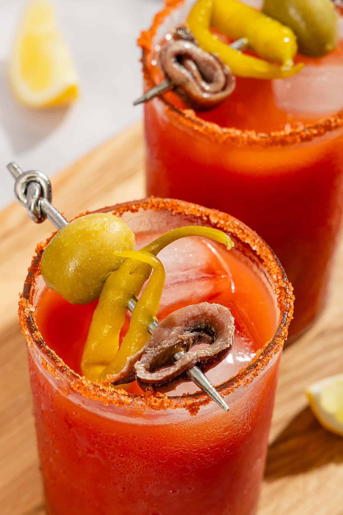 A close up of two Mediterranean bloody Marys garnished skewers of olives, pickled peppers and anchovies on a wooden cutting board.