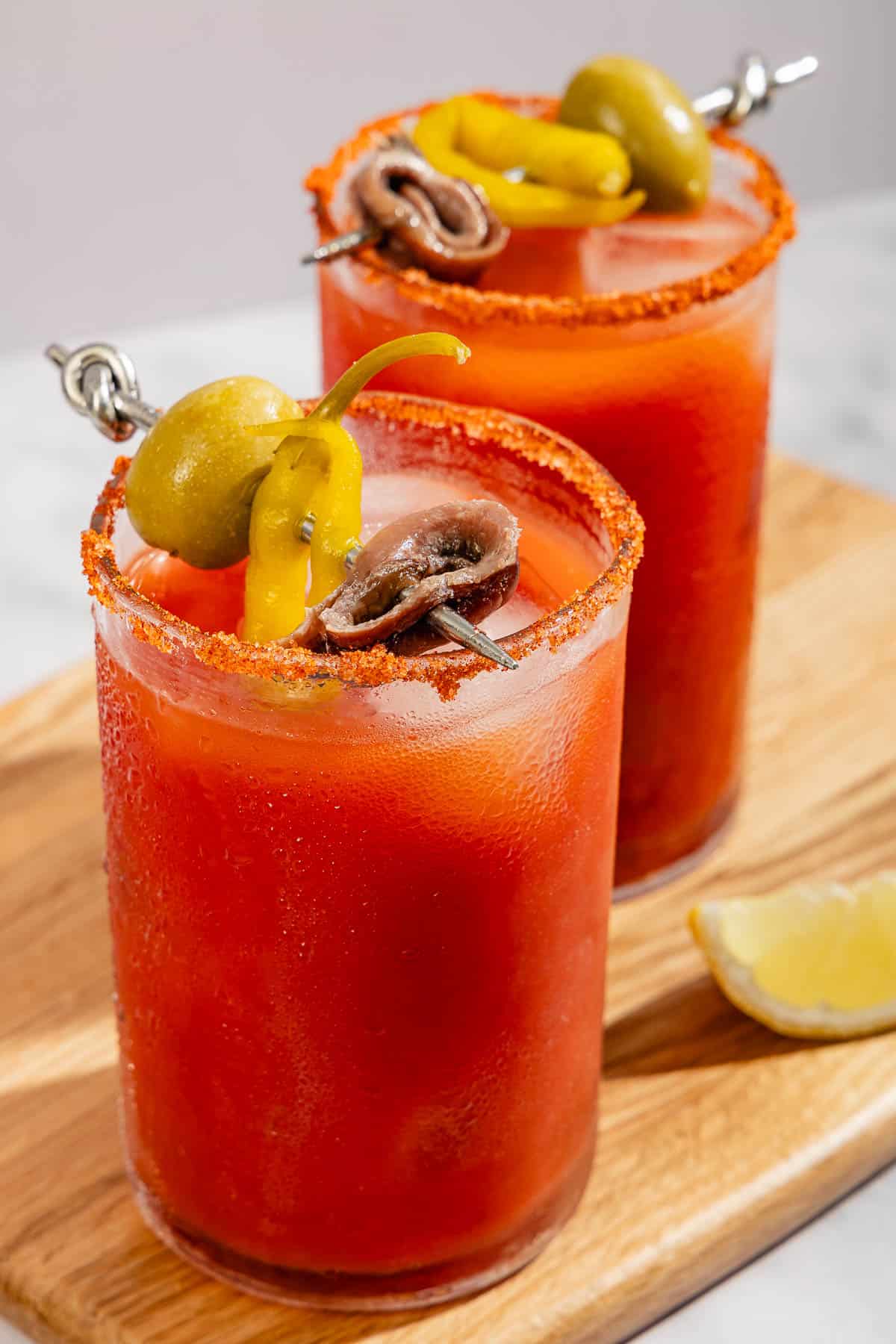 Two Mediterranean bloody Marys garnished skewers of olives, pickled peppers and anchovies on a wooden cutting board with a lemon wedge.