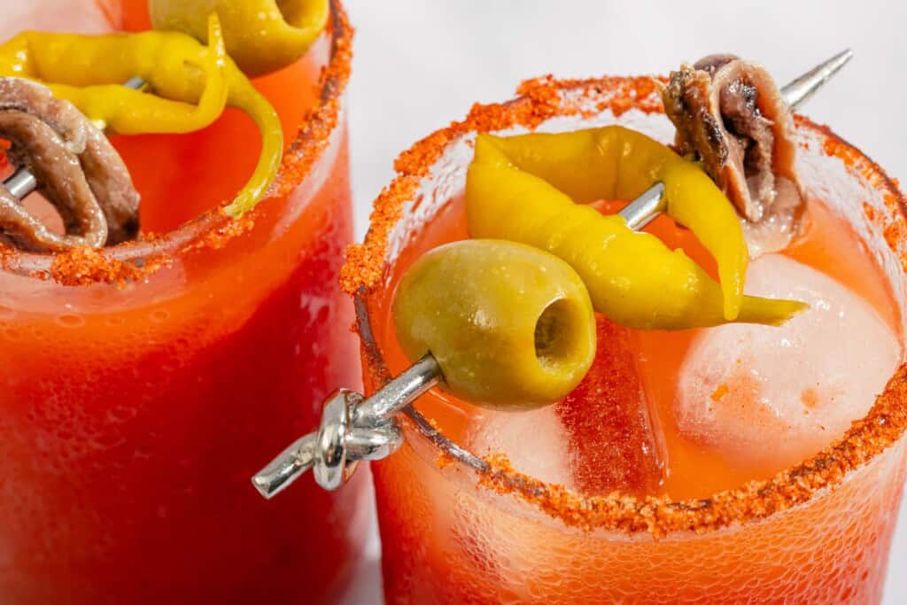 A close up of two Mediterranean bloody Marys garnished skewers of olives, pickled peppers and anchovies.