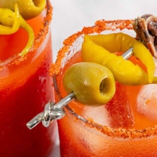 A close up of two Mediterranean bloody Marys garnished skewers of olives, pickled peppers and anchovies.