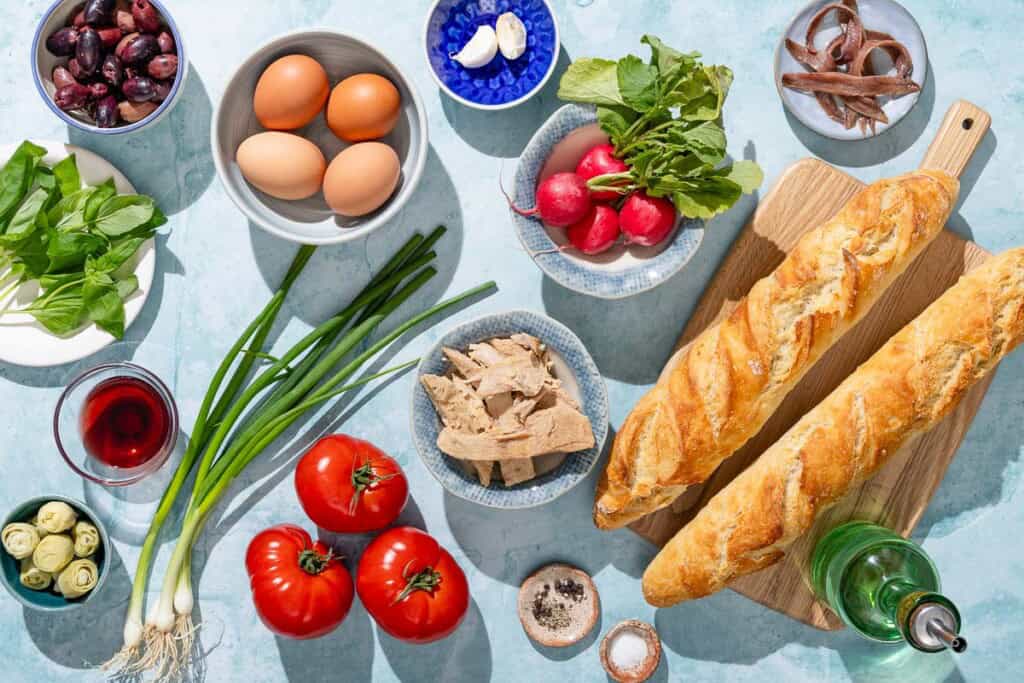 Ingredients for pan bagnat including baguettes, anchovies, radishes, eggs, kalamata olives, basil, tuna, tomatoes, green onions, artichoke hearts, red wine vinegar, salt, black pepper and olive oil.