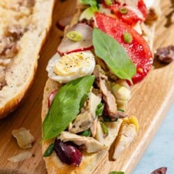 A scooped out baguette half drizzled with dressing next to a baguette half topped with vegetables, tuna and hard boiled eggs on a wooden cutting board.