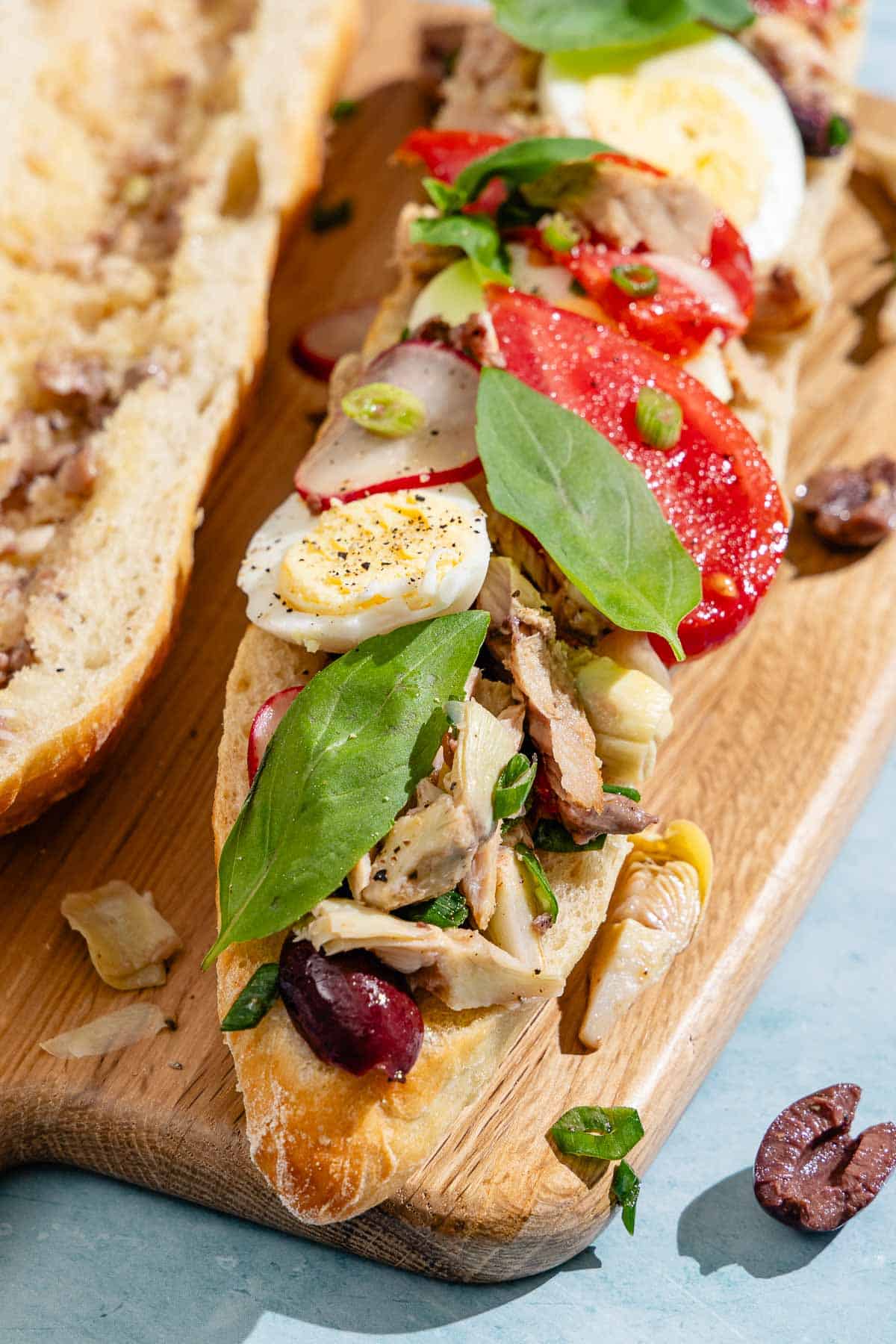 A scooped out baguette half drizzled with dressing next to a baguette half topped with vegetables, tuna and hard boiled eggs on a wooden cutting board.