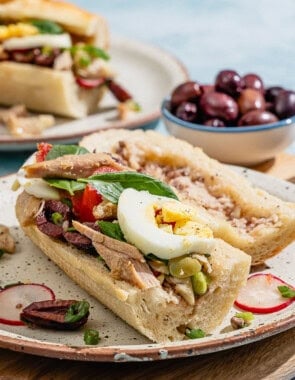 A pan bagnat with its top off sitting next to it on a plate. Next to this is another plate with a pan bagnat, two glasses of white wine, and bowls of kalamata olives and salt.