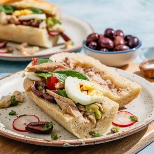 A pan bagnat with its top off sitting next to it on a plate. Next to this is another plate with a pan bagnat, two glasses of white wine, and bowls of kalamata olives and salt.