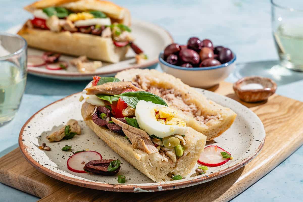 A pan bagnat with its top off sitting next to it on a plate. Next to this is another plate with a pan bagnat, two glasses of white wine, and bowls of kalamata olives and salt.