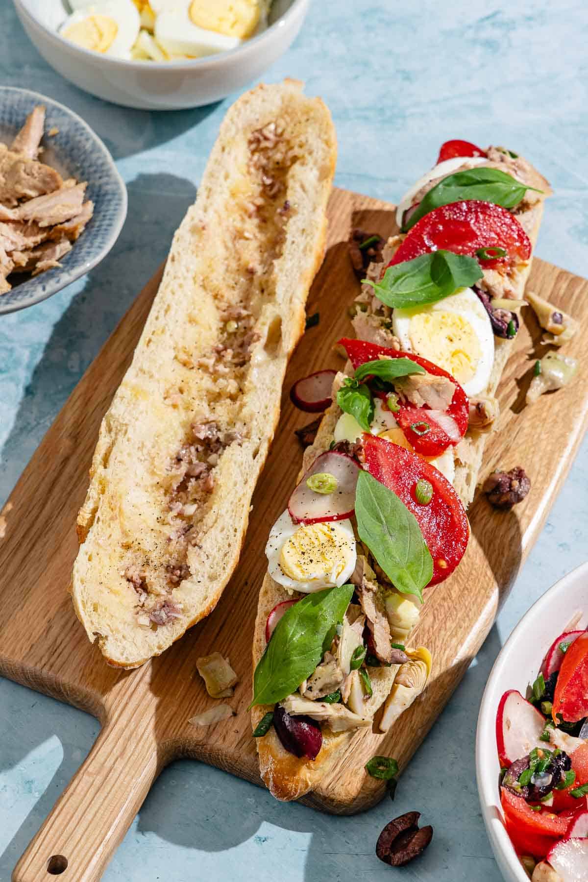 A scooped out baguette half drizzled with dressing next to a baguette half topped with vegetables, tuna and hard boiled eggs on a wooden cutting board. Next to these are bowls of the tuna, sliced hard boiled eggs and veggies.