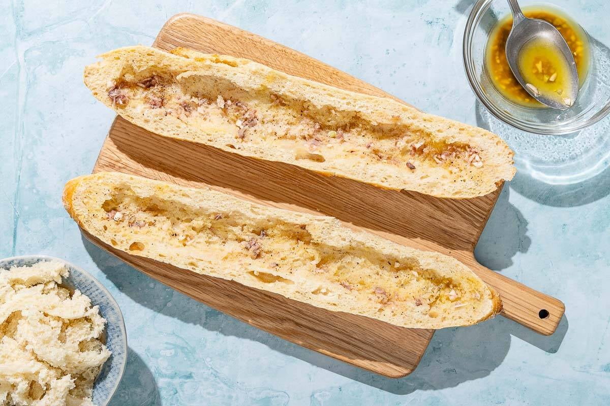 Two scooped out baguette halves drizzled with dressing on a wooden cutting board. Next to these is a bowl of the dressing with a spoon, and a bowl with the scooped our portion of the baguettes.