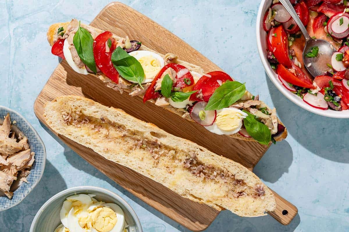 A scooped out baguette half drizzled with dressing next to a baguette half topped with vegetables, tuna and hard boiled eggs on a wooden cutting board. Next to these are bowls of the tuna, sliced hard boiled eggs and veggies.