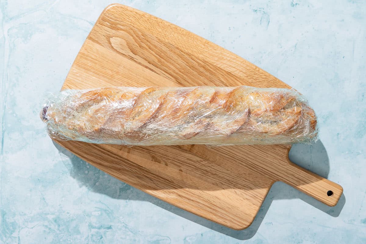 A baguette covered in plastic wrap in a wooden cutting board.