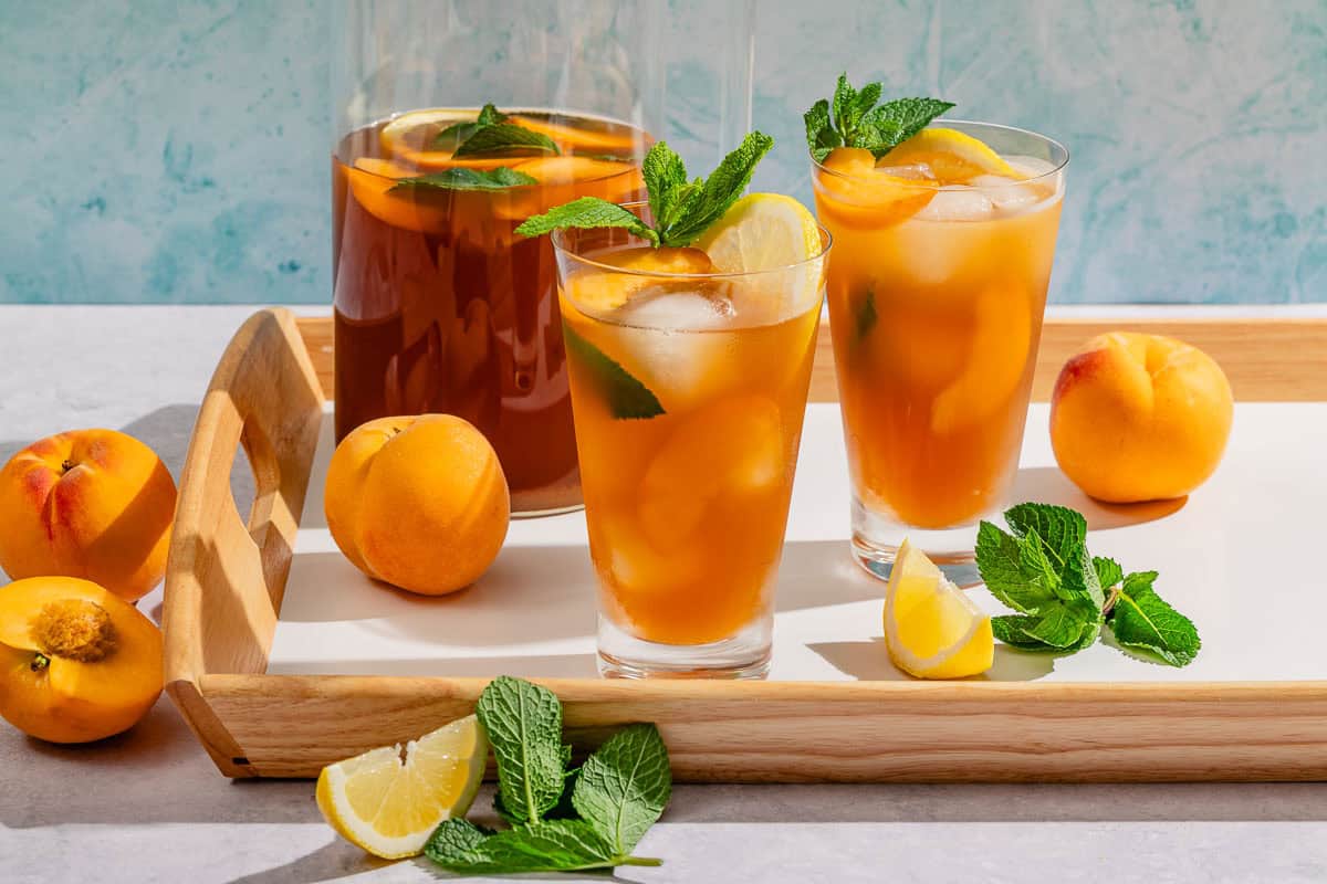 Two glasses of peach tea garnished with mint and peach and lemon wedges. Next to this is a pitcher of the tea, peaches, lemon wedges and fresh mint.