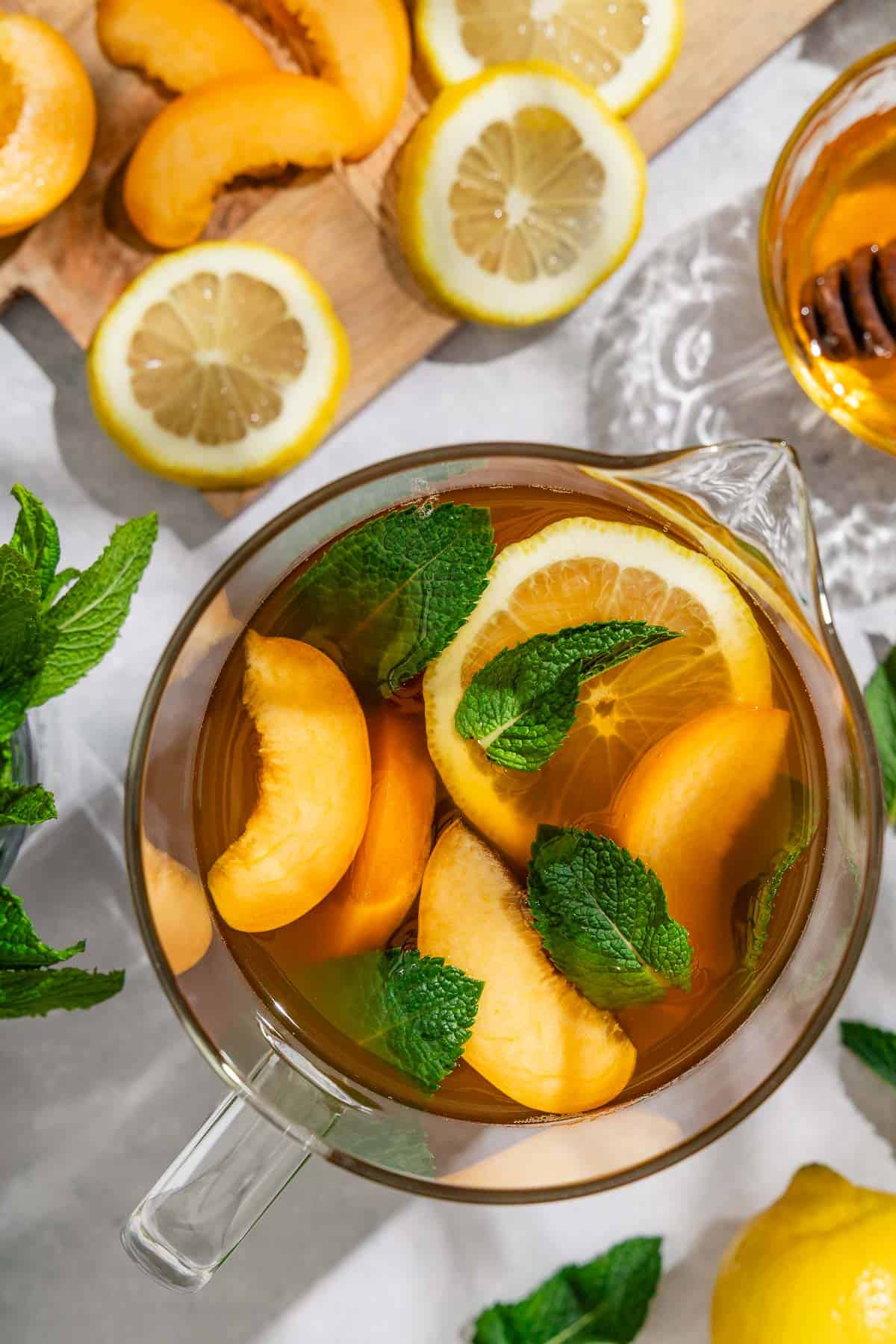 A pitcher of peach tea next to a cutting board with sliced peaches and lemon wheels and a bowl of honey with a honey dipper.