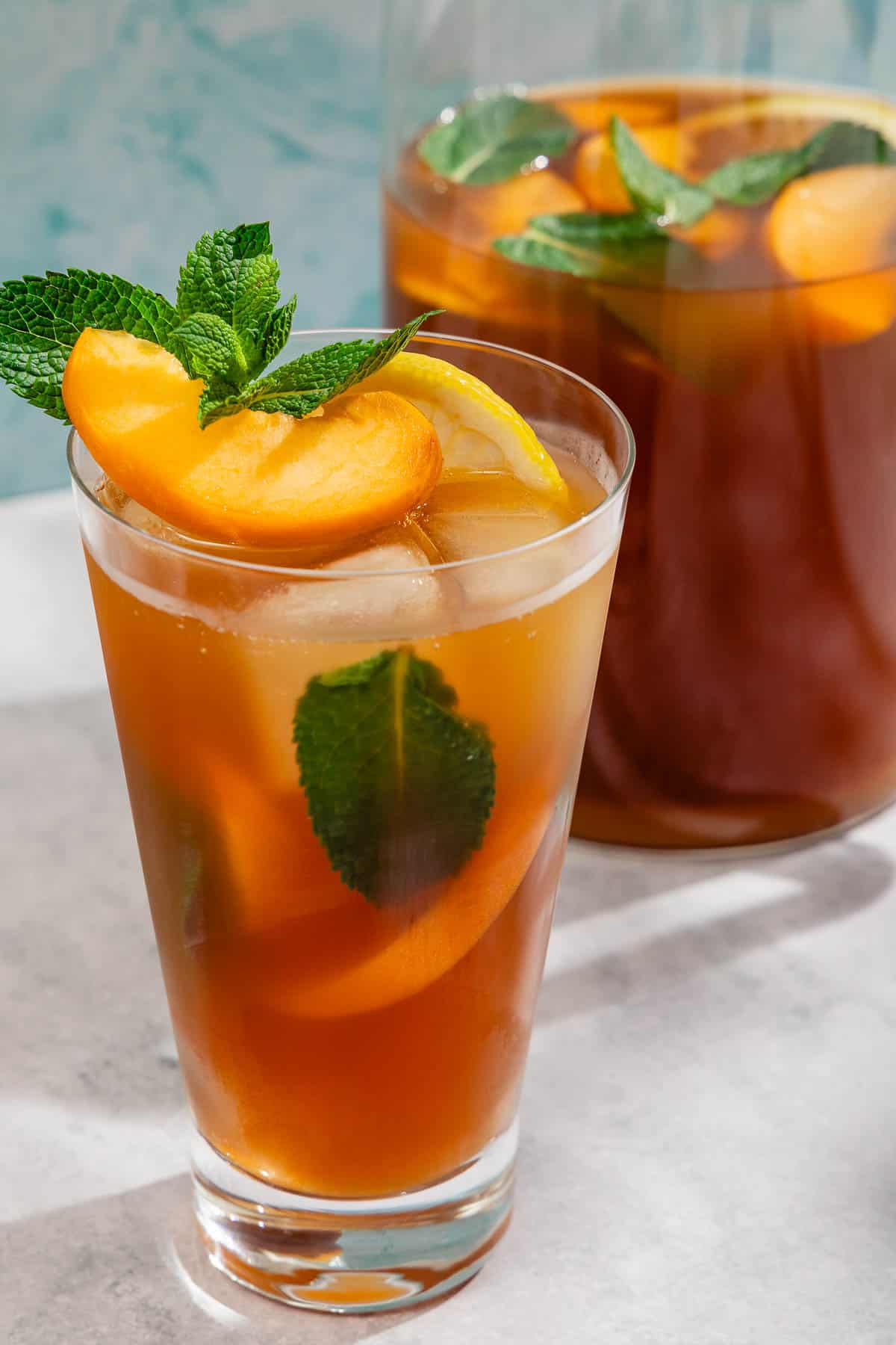 A glass of peach tea garnished with mint and peach and lemon wedges next to a pitcher of the tea.