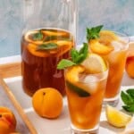 Two glasses of peach tea garnished with mint and peach and lemon wedges. Next to this is a pitcher of the tea, peaches, lemon wedges and fresh mint.