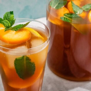 A glass of peach tea garnished with mint and peach and lemon wedges next to a pitcher of the tea.