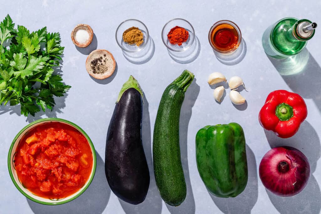 Ingredients for Spanish pisto including eggplant, zucchini, green bell pepper, red bell pepper, redo onion, garlic, parsley, olive oil, canned diced tomatoes, salt, black pepper, ground cumin, sweet paprika, and sherry vinegar.