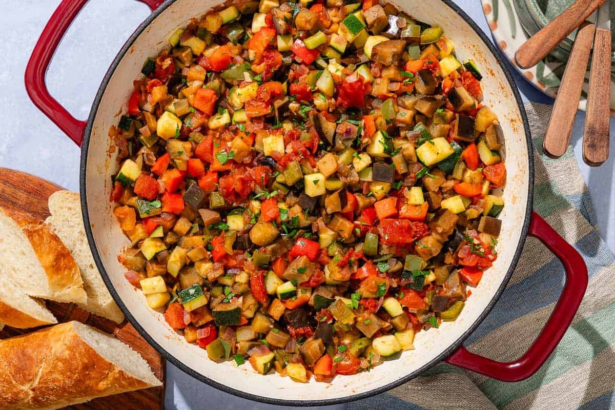 Spanish pisto in a skillet next to some slices of crusty bread and a stack of plates and utensils.