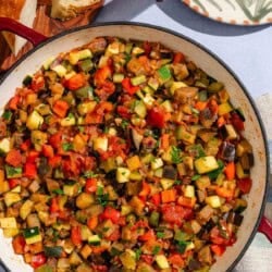 Spanish pisto in a skillet next to some slices of crusty bread and a stack of plates and utensils.