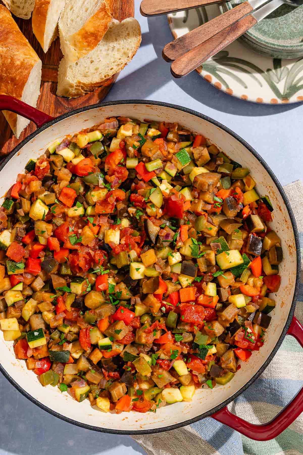 Spanish pisto in a skillet next to some slices of crusty bread and a stack of plates and utensils.