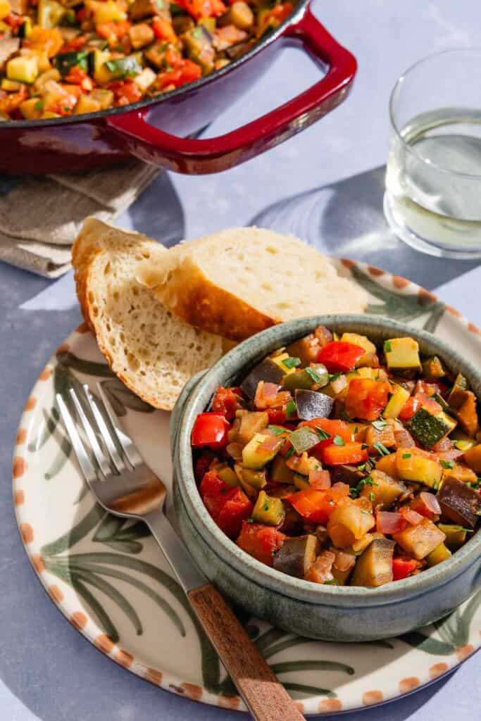 Spanish pisto in a bowl on a plate with a fork and slices of crusty bread next to a glass of white wine and the rest of the pisto in a skillet.
