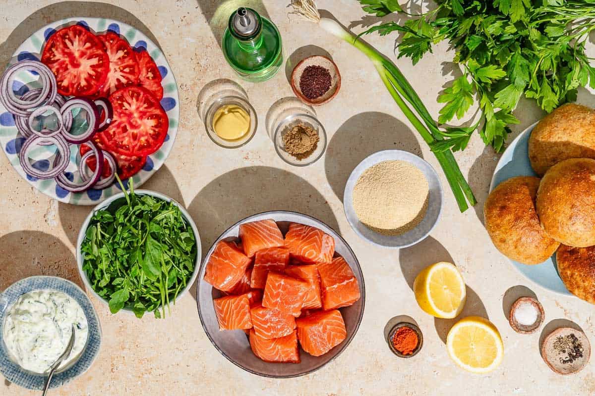 Ingredients for salmon burgers including salmon, dijon mustard green onions, parsley, ground coriander, ground sumac, sweet paprika, black pepper, salt, bread crumbs, olive oil, lemon, tzatziki sauce, arugula, red onions, tomato and buns.