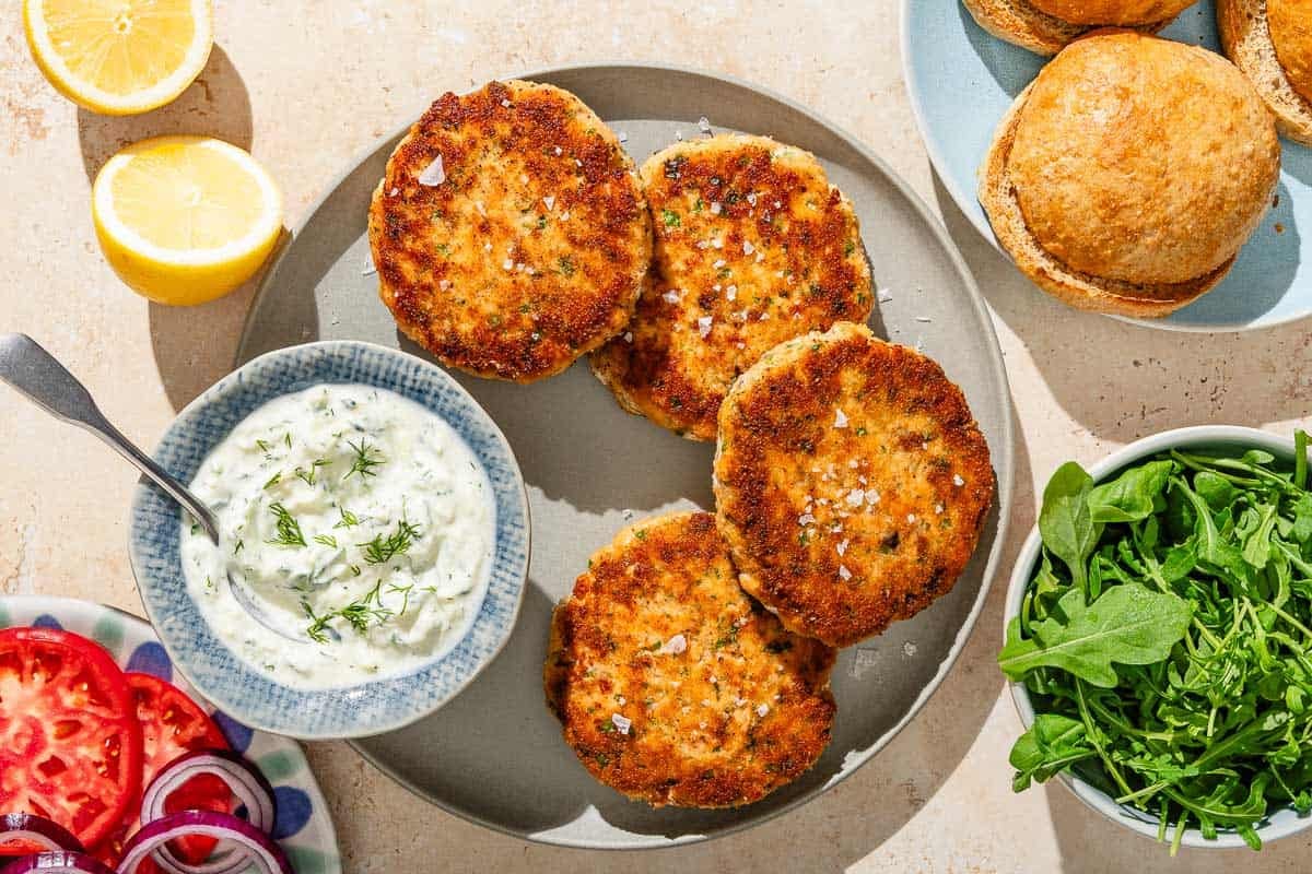 Four salmon patties on a plate with a bowl of tzatziki sauce. Surrounding this is a plate with sliced tomato and red onion, 2 lemon halves, a plate of buns, and a bowl of arugula.