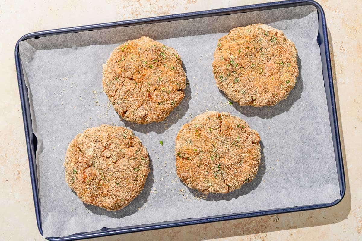 Four breaded but uncooked salmon patties on a parchment lined sheet pan.