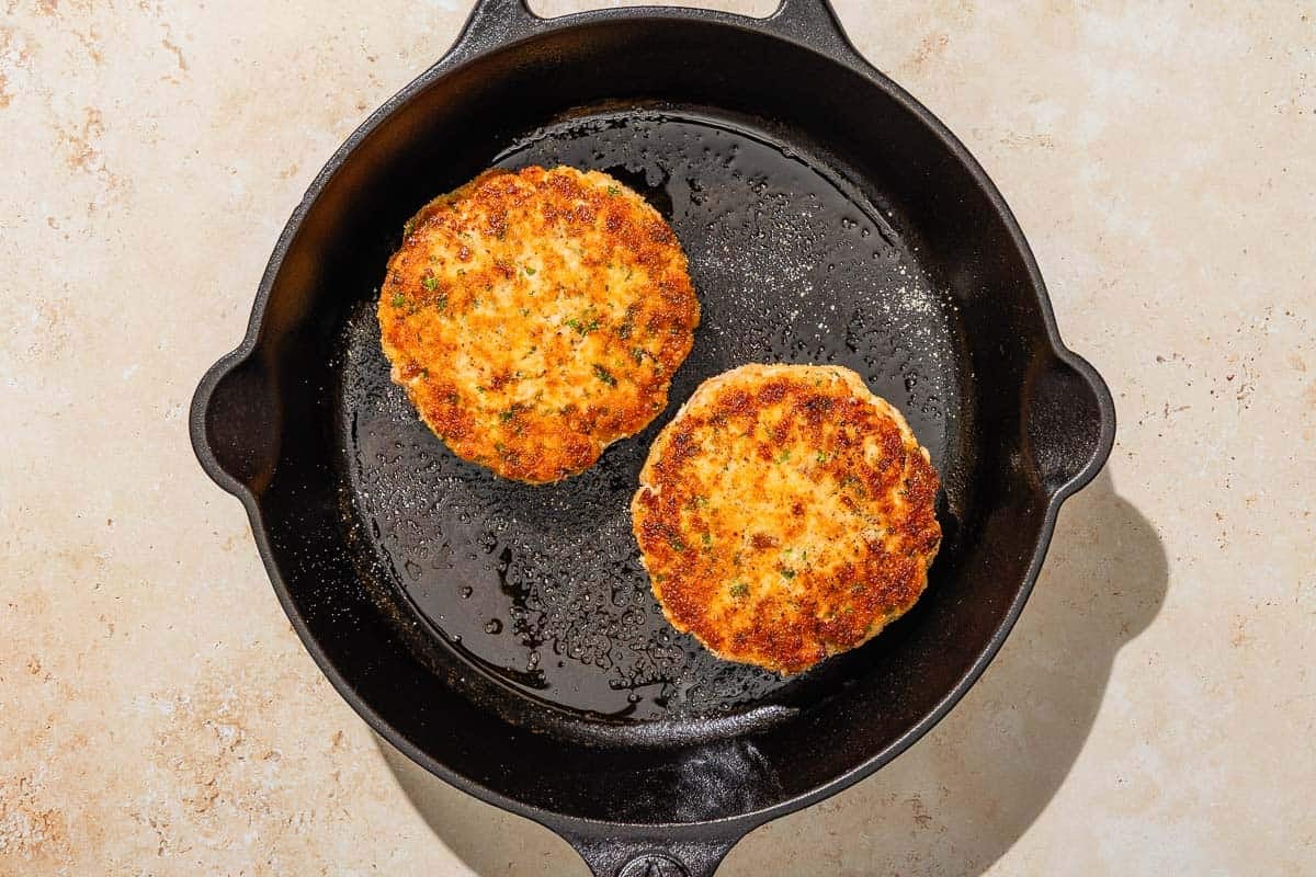 Two salmon patties being fried in a cast iron skillet.