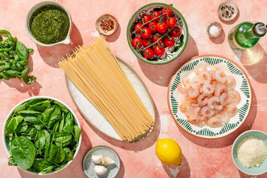 Ingredients for shrimp pesto pasta including shrimp, spaghetti, salt, black pepper, baking soda, olive oil, garlic, cherry tomatoes, pesto, red pepper flakes, baby spinach, grated Parmesan cheese and basil.