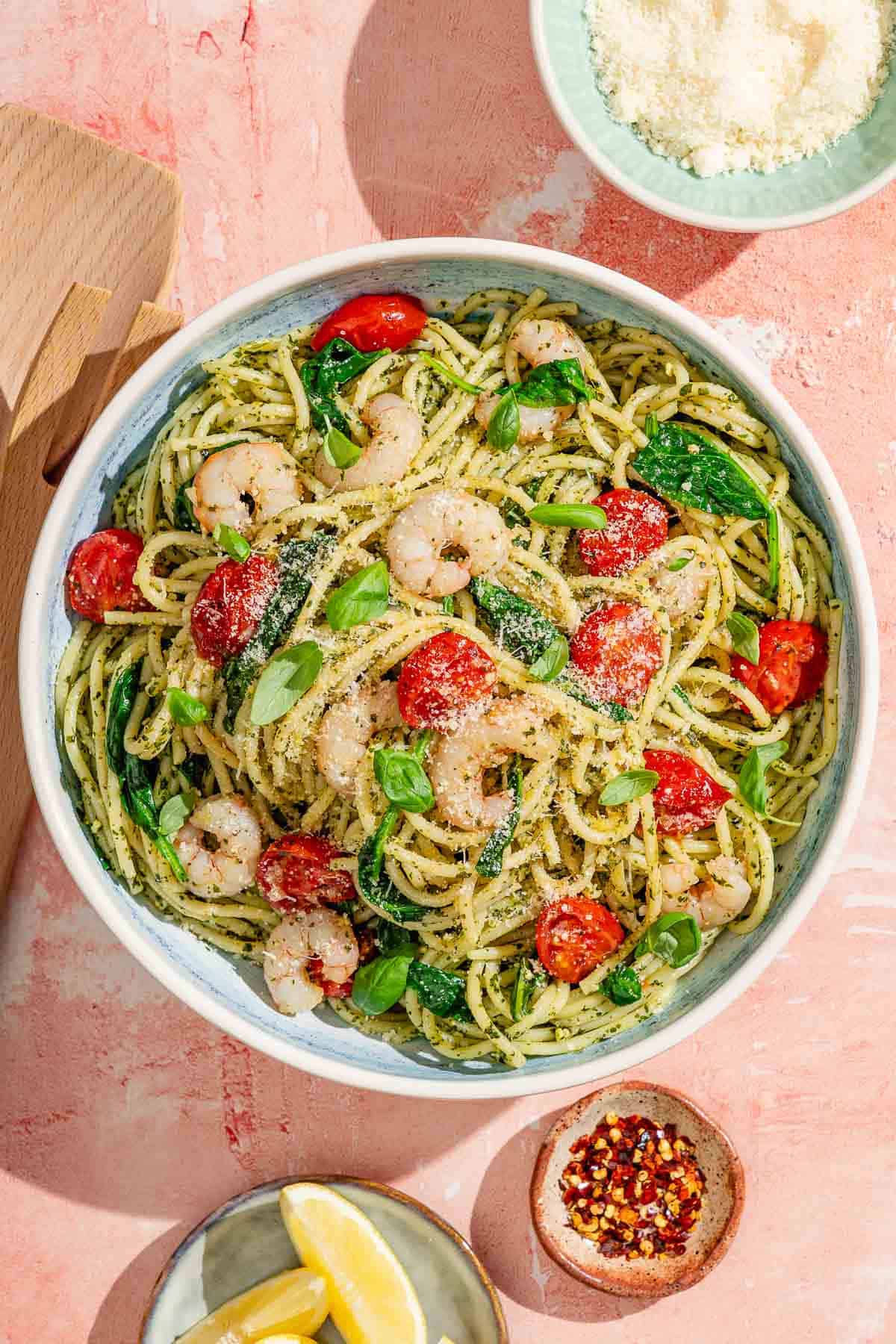 Shrimp pesto pasta in a serving bowl surrounded by a bowl of grated parmesan, a plate of lemon wedges, and a small bowl of red pepper flakes.