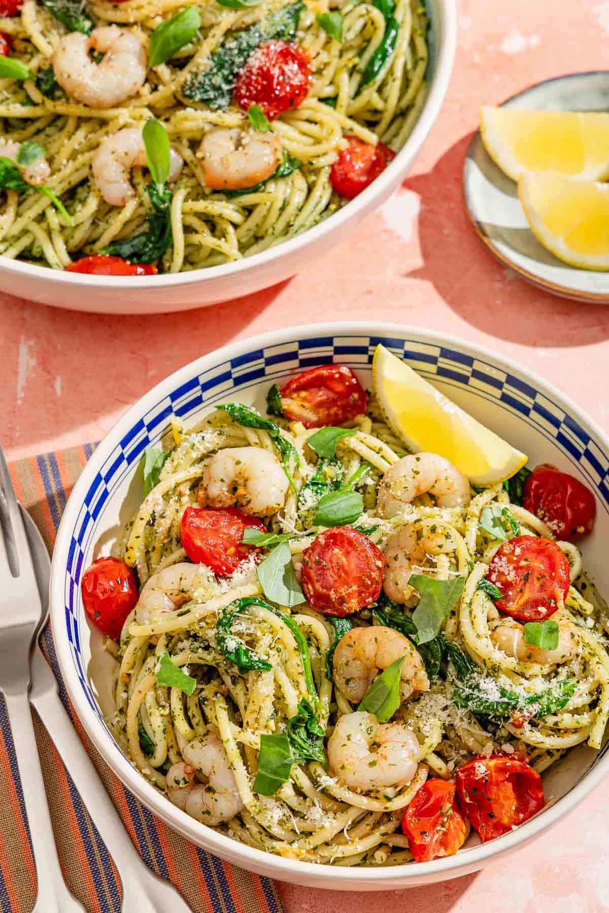 A serving of the shrimp pesto pasta in a bowl with a lemon wedge. Next to this is a fork and spoon, a plate of lemon wedges, and a serving bowl with the rest of the pasta dish.