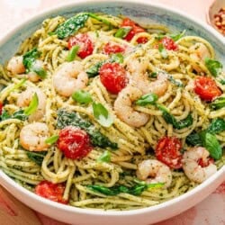 Shrimp pesto pasta in a serving bowl surrounded by a bowl of grated parmesan, a plate of lemon wedges, and a small bowl of red pepper flakes.