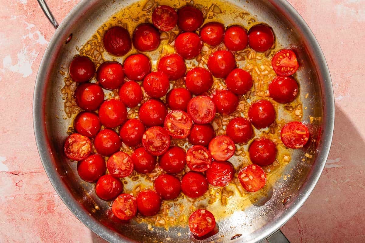 The cherry tomatoes for the shrimp pesto pasta being cooked with garlic in olive oil in a tall-sided skillet.