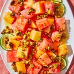 Cubed watermelon, cantaloupe and lime wheels on a platter topped with a honey and brown sugar dressing and pistachios.