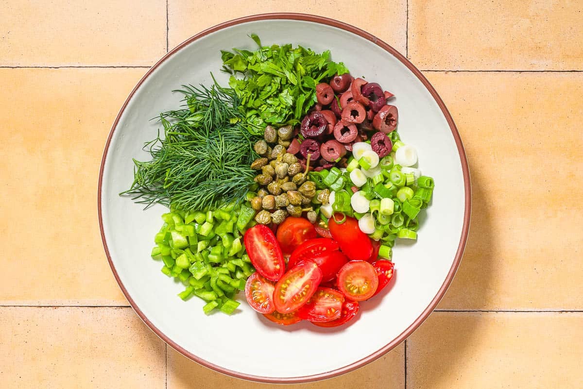 A bowl of the chopped parsley, dill, green pepper, and green onions, slices cherry tomatoes and kalamata olives, and capers in a bowl before being mixed together.