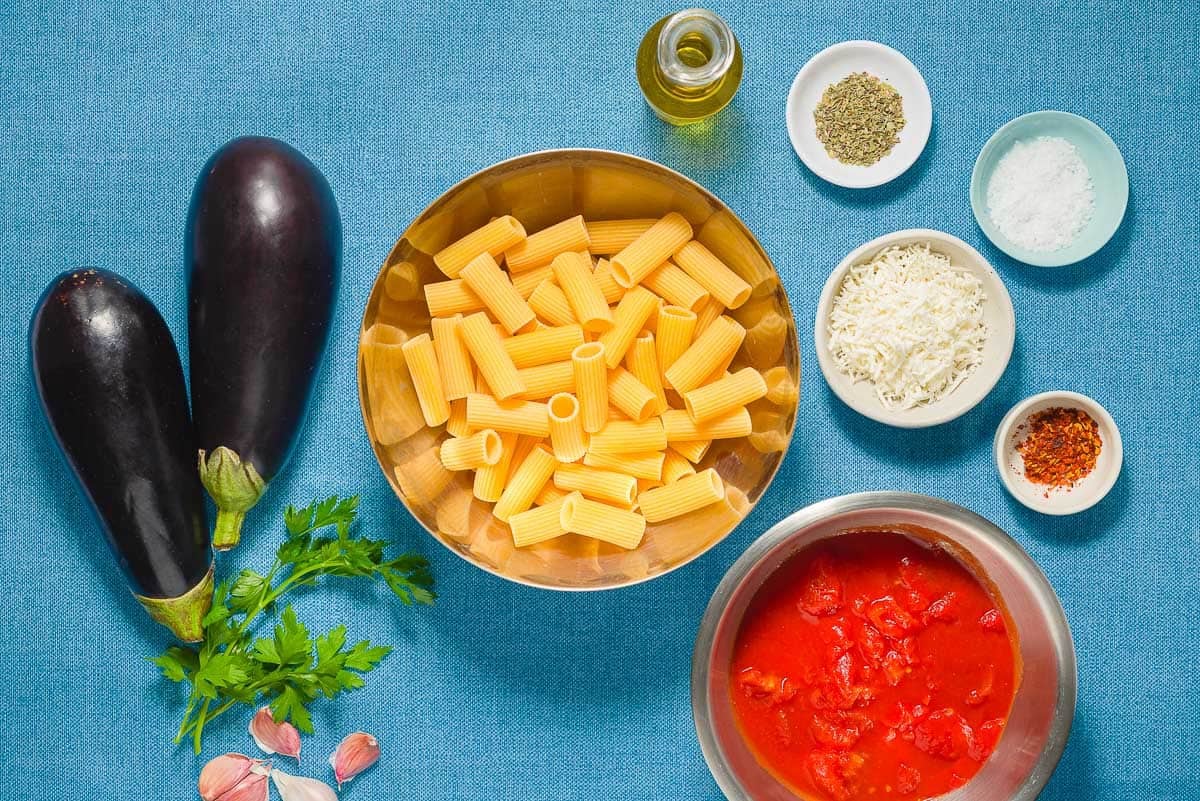 ingredients for pasta alla norma including eggplants, salt, olive oil, garlic, red pepper flakes, canned diced tomatoes, dried oregano, rigatoni pasta, parsley and grated ricotta salata.